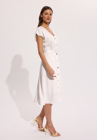 Kobieta w białej, zapinanej na guziki sukience midi z krótkimi rękawami i paskiem, w połączeniu ze złotymi sandałami na wysokim obcasie, stojąca bokiem.