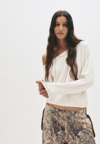 Weißer, locker sitzender, schulterfreier Pullover mit langen Ärmeln. Kombiniert mit gemusterten, mehrfarbigen Hosen in erdigen Tönen und einem gesteckten Design.