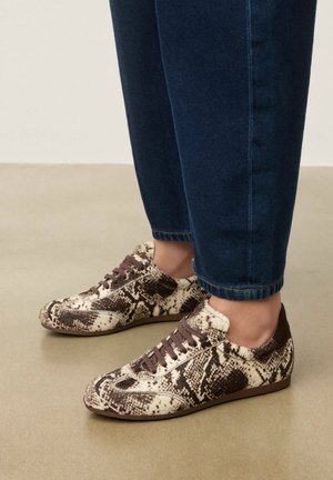 Bruine en crème sneakers met slangenprint, gedragen met donkerblauwe spijkerbroek op een beige vloer.