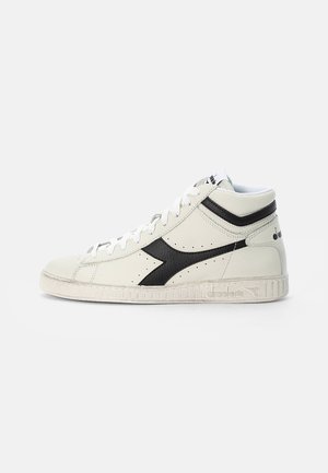 Baskets montantes blanches avec détail de bande noire, lacets blancs et semelle texturée, présentées sur un fond gris clair uni.