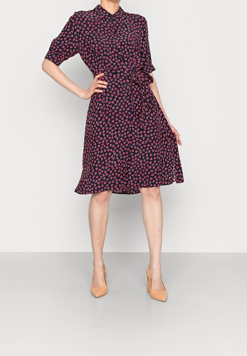 Femme portant une robe noire mi-longue avec des motifs de cœurs roses et des talons hauts couleur chair, debout avec une main sur la hanche.