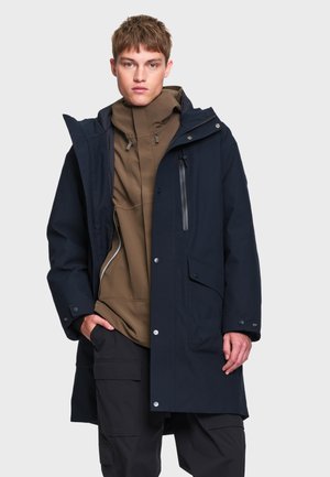Jeune homme portant un sweat à capuche marron sous un imperméable bleu marine avec des détails de fermeture éclair et de boutons, posant avec une main dans la poche sur un fond gris.
