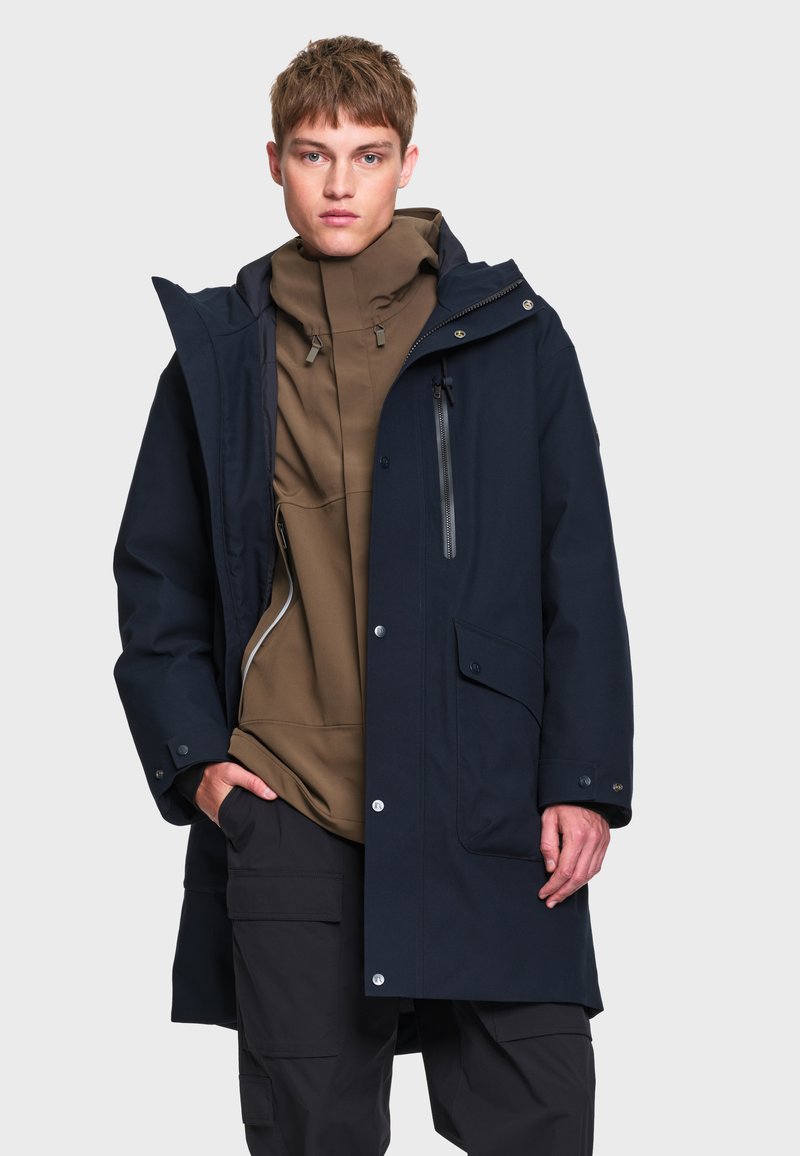 Jeune homme portant un sweat à capuche marron sous un imperméable bleu marine avec des détails de fermeture éclair et de boutons, posant avec une main dans la poche sur un fond gris.