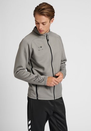 Grijze sportjack met een hoge kraag, volledige rits aan de voorkant en zwarte accenten. Voorzien van een logo en een chevronontwerp op de mouwen.