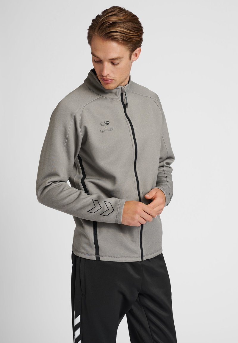 Giacca sportiva grigia con colletto alto, zip frontale completa e strisce nere di accento. Presenta un logo e un design a chevron sulle maniche.
