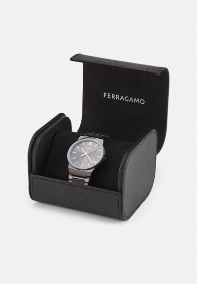 Black watch in a black textured case. Watch features a dark dial, silver accents, round face, and metallic band. Brand name visible on case.