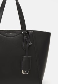 Svart skinnveske med en strukturert design, som har to håndtak, et avtakbart merke og tonede sting. Merkevaren er preget foran.