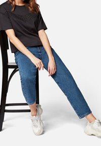 Černé krátké tričko s texturovaným, vzorovaným předním dílem, kombinované s modrými džíny a bílými masivními teniskami.