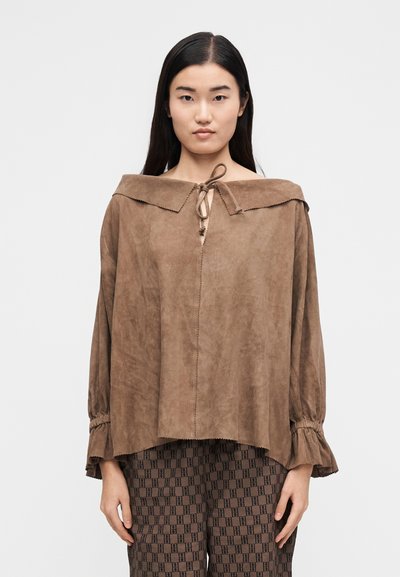 Γυναίκα με μακριά σκούρα μαλλιά φορεί ένα χαλαρό καφέ σουέτ τοπ με φαρδιά γιακά δεμένο στον λαιμό και καφέ παντελόνι με σχέδια, στέκεται μπροστά σε λευκό φόντο.