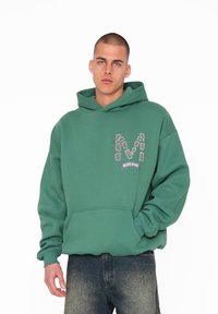 Grøn hættebluse lavet af blødt stof, med en frontlomme og et grafisk "M" logo. Designet med ribbede manchetter og nederkant.