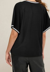 Una donna dai lunghi capelli è vista di spalle, indossa un top nero con bordi bianchi e pantaloni neri, su uno sfondo neutro.