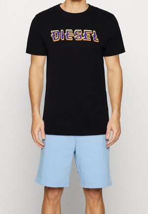 Homme portant un t-shirt noir à manches courtes avec le texte coloré "DIESEL" et un short bleu clair jusqu'aux genoux, se tenant contre un fond uni.