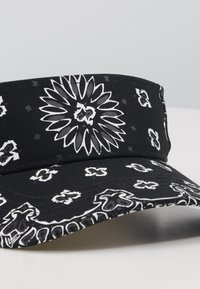 Svart caps med hvite blomster- og geometriske mønstre, med en strukturert visir og teksturert stoff. Detaljer inkluderer kontraststing.