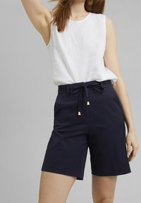 Top sin mangas blanco de lino combinado con pantalones cortos azul marino con cintura ajustable y puntas decoradas en dorado. Textura suave en general.