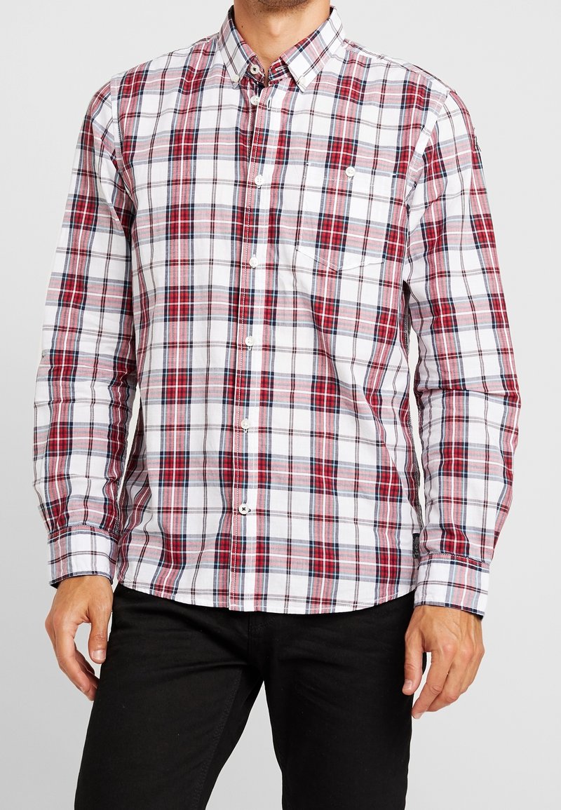 Uomo che indossa una camicia a quadri rossa, bianca e nera con maniche lunghe, bottoni e taschino sul petto, e pantaloni neri, in piedi contro uno sfondo semplice.