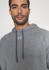 Sweat-shirt à capuche gris avec une poche kangourou, des cordons et un petit logo blanc sur la poitrine. Texture douce et coupe décontractée.