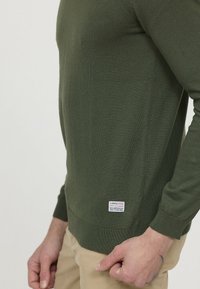 Pull en tricot vert avec une coupe ajustée, des poignets et un ourlet côtelés. Il présente une étiquette tissée en bas. Texture lisse, manches longues.