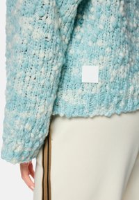 Strickpullover in helltürkis und Weiß. Grober, strukturierter Muster mit einem gerippten Saum. Verfügt über ein quadratisches Logo-Patch an der Seite.
