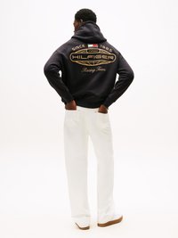 Sudadera azul marino con logo dorado en la espalda, que presenta el texto "HILFIGER Racing Team" y "DESDE 1985", combinada con pantalones blancos holgados y zapatos.