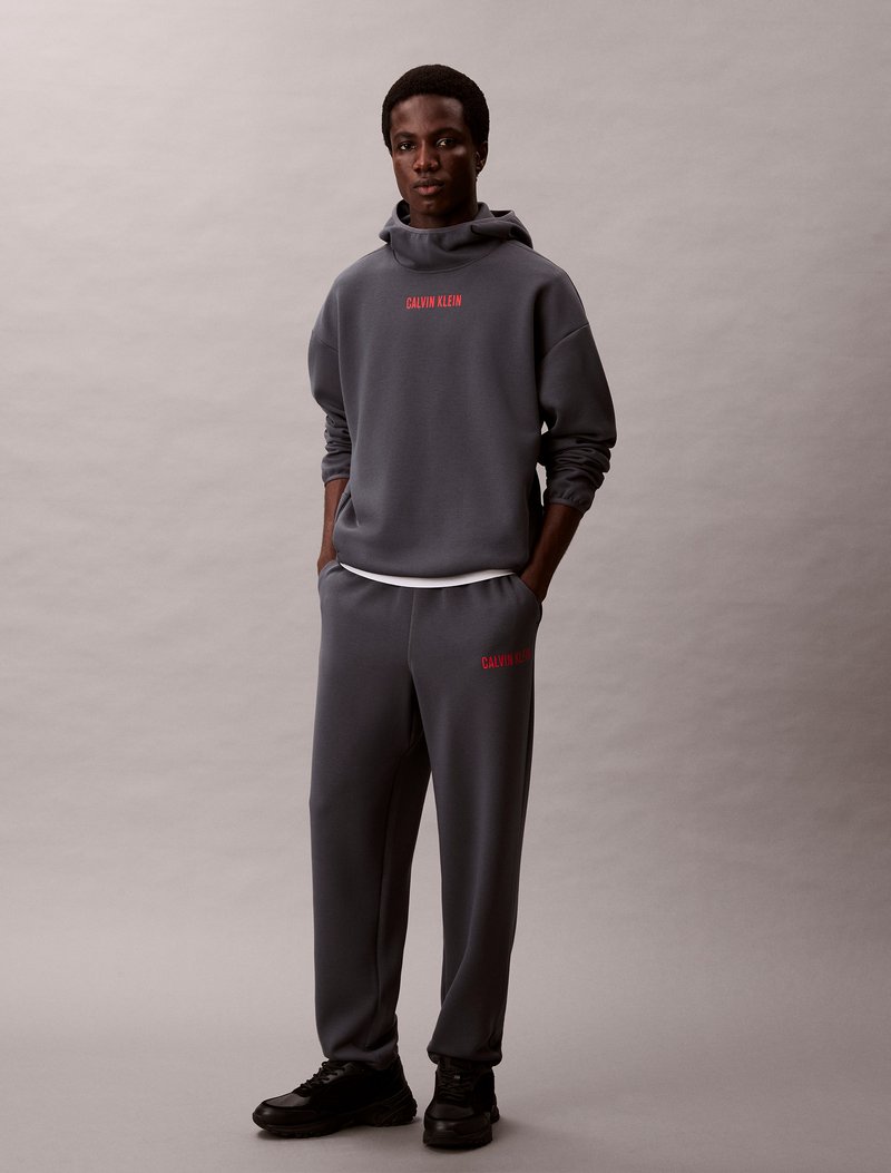 Szara bawełniana bluza z kapturem i pasujące dresowe spodnie z czerwonym logo "Calvin Klein". Posiada ściągacz na kapturze i luźny krój. Czarnie trampki dopełniają cały strój.