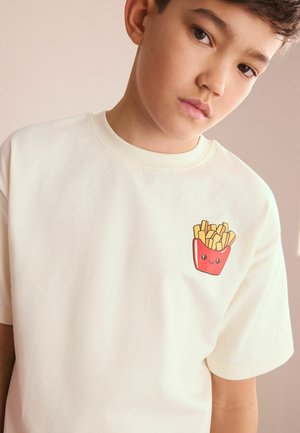 Fiú, aki fehér pólót visel, amin kis piros sültkrumpli rajzfilmfigura van a mellkason, egy egyszínű háttér előtt áll.