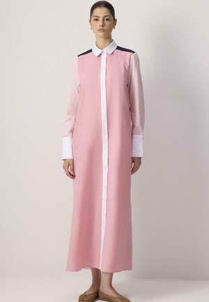 Femme portant une longue robe rose boutonnée avec un col et des poignets blancs, des manches rayées et des chaussures plates marron, debout devant un fond uni.