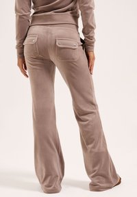 Lichtbruine uitlopende broek van zacht, glad stof, met twee achterzakken met drukknoopsluitingen. De tailleband is elastisch.