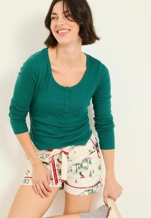 Eine Frau lächelt, trägt ein grünes Longsleeve-Shirt und cremefarbene Shorts mit Ski-Motiven und hält einen Hausschuh in der Hand, während sie an einem Fuß einen grauen Socken trägt.