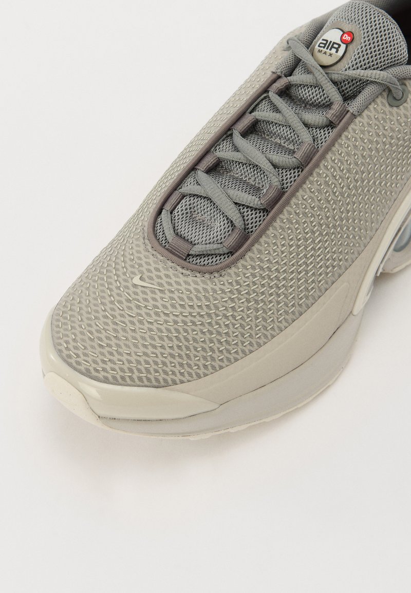 Zapatilla gris claro con parte superior de malla texturada, cordones grises y una unidad de aire visible en el talón. Presenta una punta redondeada y un branding mínimo.