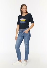 Marineblauwe T-shirt met korte mouwen en een grafisch tekstontwerp; gecombineerd met lichtblauwe slim-fit jeans met verweringsdetails en witte sneakers.