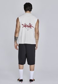 Biała koszulka bez rękawów z czerwonym tekstem graficznym "SikSilk" na plecach, w połączeniu z czarnymi szortami do kolan i białymi skarpetkami, noszona z czarnymi butami.