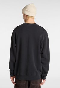 Sort bomulds sweatshirts med afslappet pasform, ribbede håndled og kant. Bæres lagdelt over brune bukser. En lys beige strikket hue bæres på hovedet.