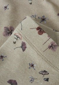 Beżowa tkanina złożona, z przeszytymi brzegami, ozdobiona małymi, fioletowymi i brązowymi wzorami kwiatowymi oraz motylami rozrzuconymi po powierzchni.