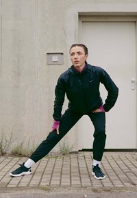 Kobieta w czarnym stroju sportowym rozciąga nogi na świeżym powietrzu na wyłożonej kostką powierzchni w pobliżu jasnych drzwi i betonowej ściany.