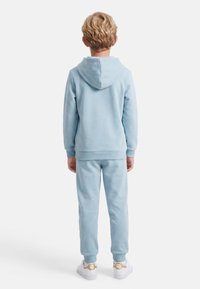 Παιδί με ξανθά μαλλιά που φοράει ανοιχτό μπλε φούτερ με κουκούλα και αντίστοιχο παντελόνι φόρμας, στέκεται πλάτη σε λευκό φόντο.