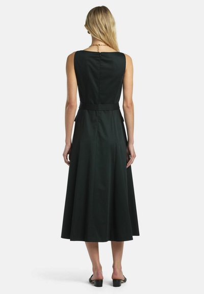 Frau mit langen blonden Haaren, die ein ärmelloses schwarzes Midikleid, einen dünnen Gürtel, schwarze Schuhe mit niedrigem Absatz und eine goldene Halskette trägt, mit dem Rücken zur Kamera.