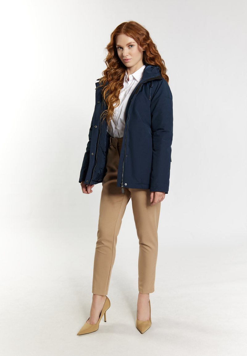 Navy-blauer Puffer-Jacke mit Kapuze, Reißverschlüssen und Knöpfen, kombiniert mit beigen, eng anliegenden Hosen und hellbraunen Absätzen. Geschmeidige Stofftextur dargestellt.