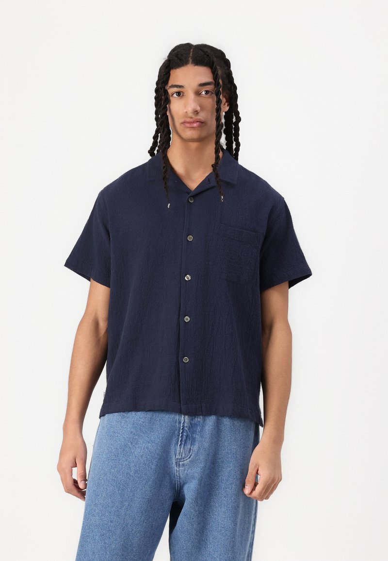 Navy short-sleeve button-up shirt with a textured finish, single chest pocket, and round collar. Worn with light blue denim jeans.