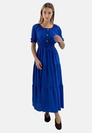 Niebieska maxi sukienka z bufiastymi rękawami, marszczonym pasem i detalami z guzikami. Spódnica ma warstwowe falbany. Modelka nosi czarne buty.