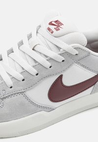 Valkoinen Nike SB Force 58 -lenkkari, jossa on vaaleanharmaita mokkanahkaosia ja viininpunainen Nike-swoosh-logo sivulla ja kielen kohdalla.