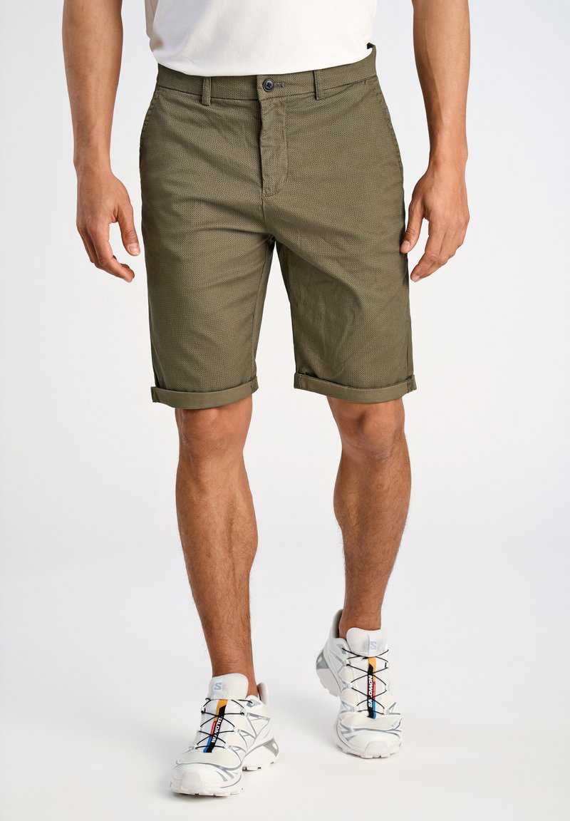 Olijfgroene korte broek met een structuurpatroon, opgerolde manchetten en een voorste knoopsluiting, gecombineerd met witte sneakers met gekleurde veters.