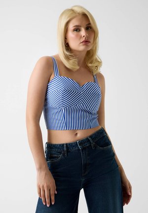 Femme blonde portant un crop top rayé bleu et blanc et un jean taille haute bleu foncé, debout devant un fond clair uni.