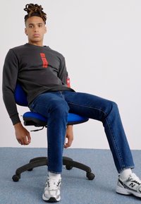 Šedá mikina s dlhým rukávom a červeným logom, modré džínsy a šedé tenisky. Sedí v čiernej ergonomickej stoličke na modrom koberci.