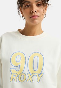 White sweatshirt featuring a large yellow and blue "90 Roxy" graphic on the front, round neckline, and textured fabric.