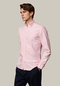 Mladý muž s hnedými vlasmi, oblečený v svetlo ružovej košeli na gombíky a tmavých nohaviciach, stojaci s rukami v vreckách na jednoduchom pozadí.