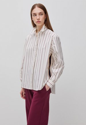 Kobieta nosząca luźną białą koszulę zapinaną na guziki z pionowymi paskami i burgundowe spodnie, stojąca z jedną ręką w kieszeni, patrząca na bok.