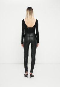 Črn dolg rokav bodysuit z majhnimi okrasnimi elementi, v kombinaciji s prilegajočimi črnimi hlačami iz umetnega usnja. Model nosi sandale na visoki peti z dodatki.