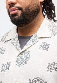 Κοντινό πλάνο ενός άνδρα με γένια που φοράει μία κρεμ πουκάμισο με κουμπιά, διακοσμημένο με σκοτεινά γκρι γεωμετρικά λουλουδένια μοτίβα, και γκρι εσώρουχο.