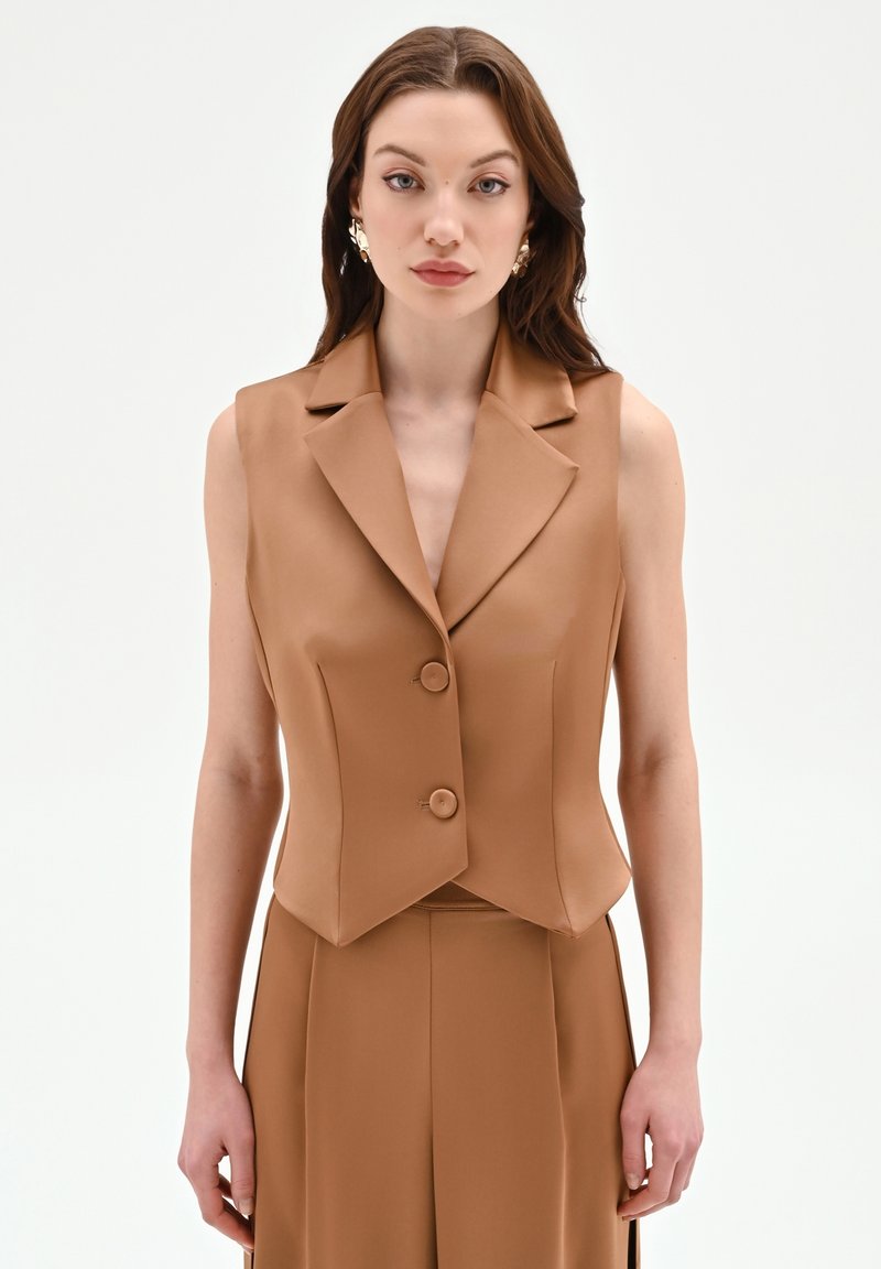 Donna con capelli castani lunghi che indossa un gilet senza maniche color sabbia con bottoni e pantaloni abbinati, in piedi davanti a uno sfondo bianco uniforme.