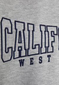 Sudadera gris con un destacado logo bordado en azul marino que dice "CALIF" y "WEST" debajo, mostrando una textura suave y un diseño informal.